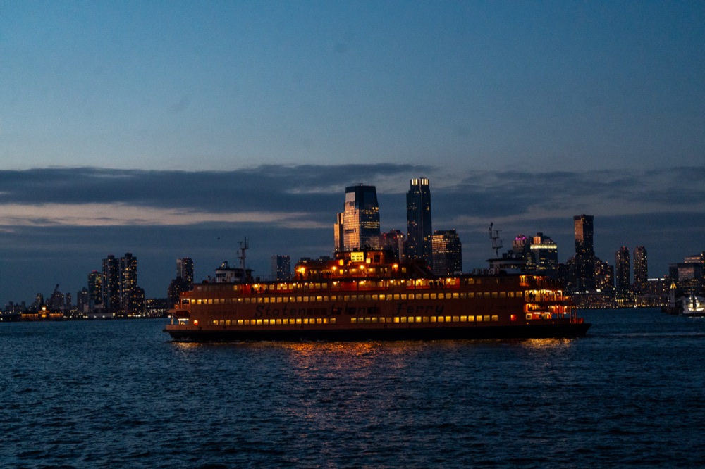 A glowing Staten Island Ferry crossing the water in front of a distant skyline.