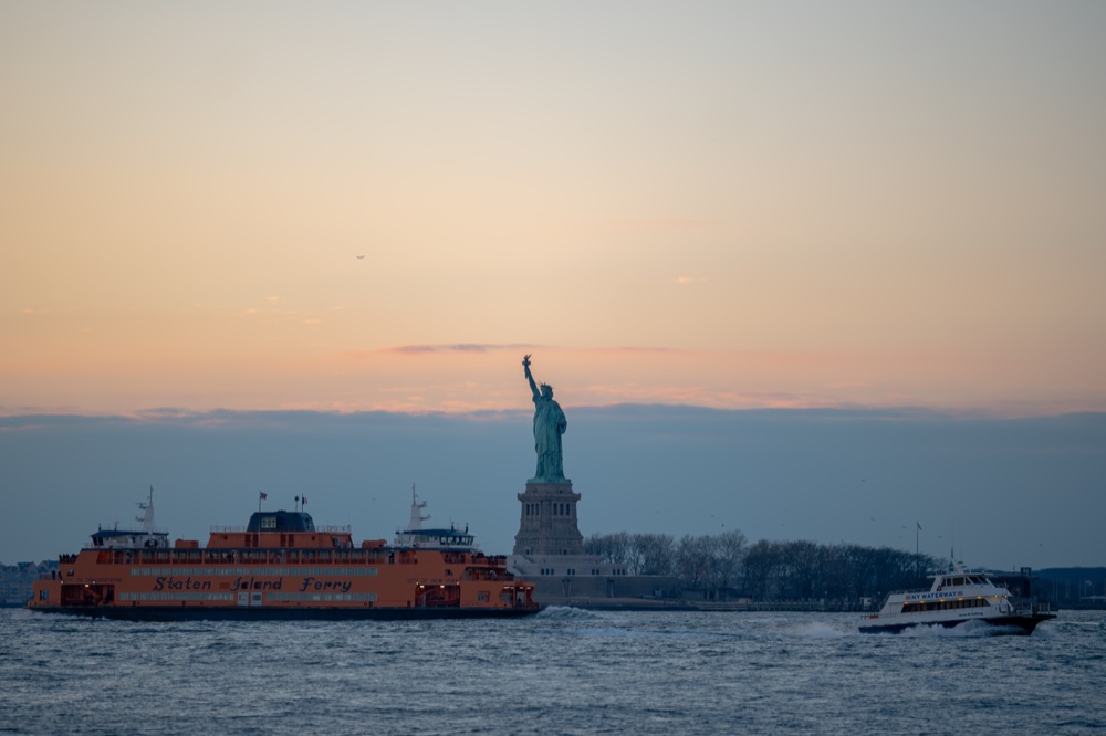 The Statue of Liberty and ferries on the water beneath a soft evening sky.