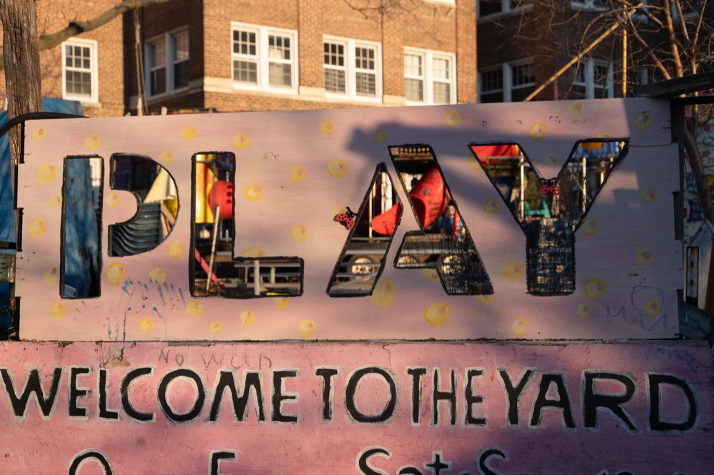 Afternoon light across a painted wooden PLAY sign beside a playground.